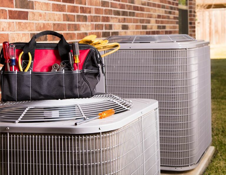 A central air conditioning unit sits outside a brick building, with a toolbox on top of the unit, suggesting maintenance or repair.